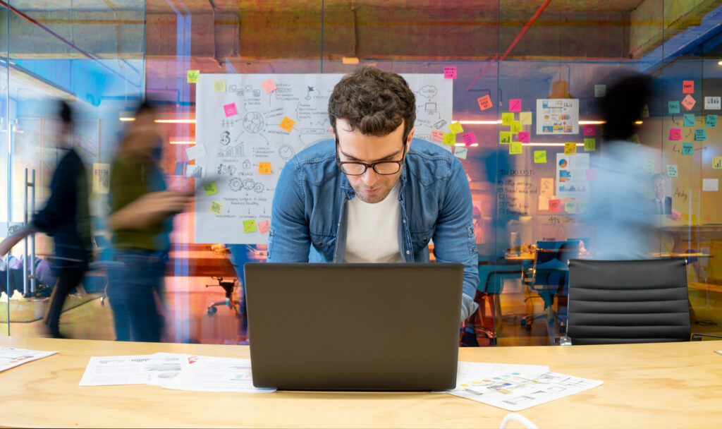 Latin American man working at a creative office using his computer and people moving at the background - place of work concepts